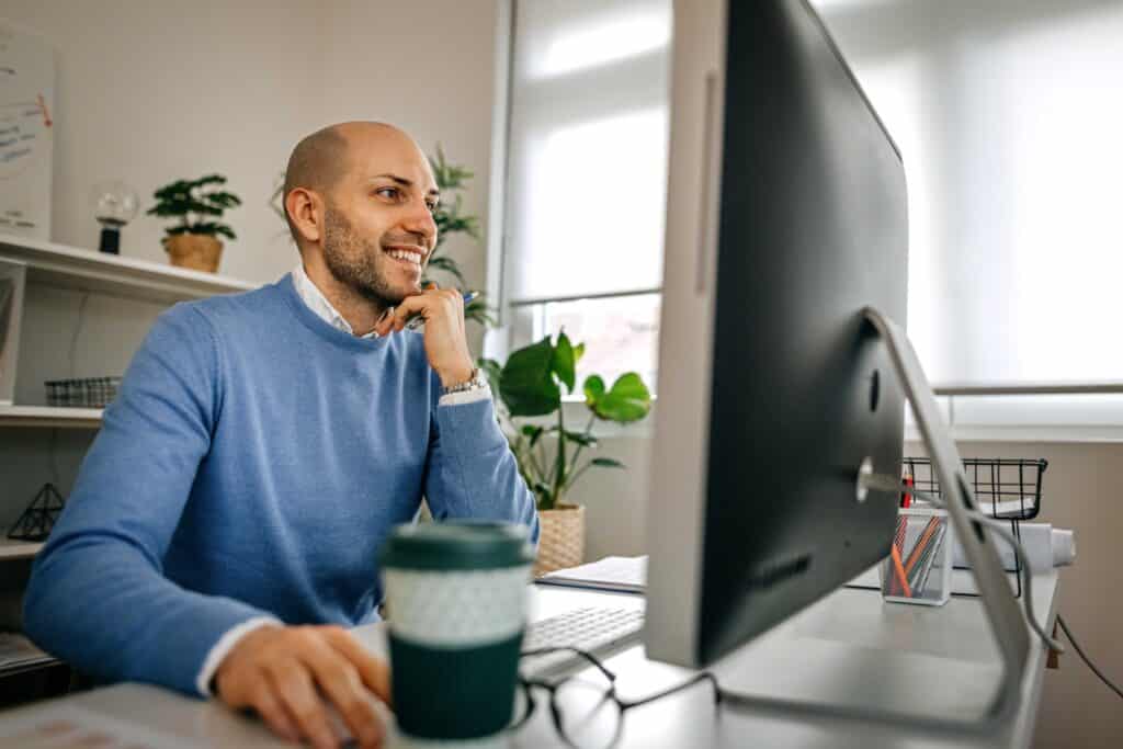 Male in blue top sits with a coffee at his desktop computer, smiling,