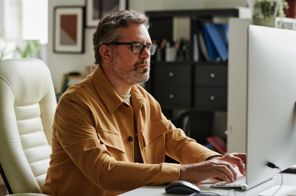 Male in yellow jacket and glasses sits typing at his computer